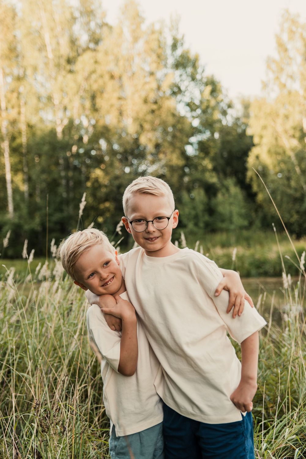 Two children in white shirts stand together posing outdoors in a grassy field with trees in the background.