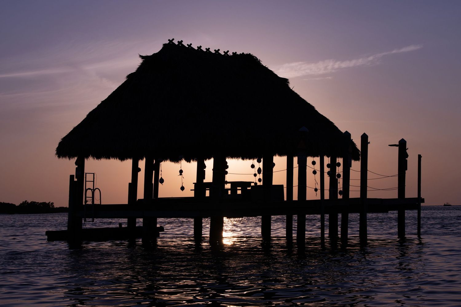 Silhouettes of stilt houses and piers stand against a dramatic purple sunset over calm ocean waters.