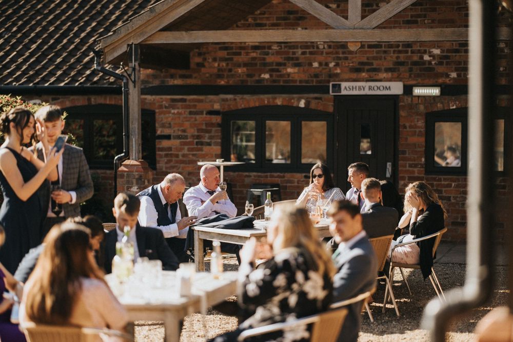 Wedding guests are gathered at tables under a rustic brick building at Hall Farm - image captured by Grimsby photographers.