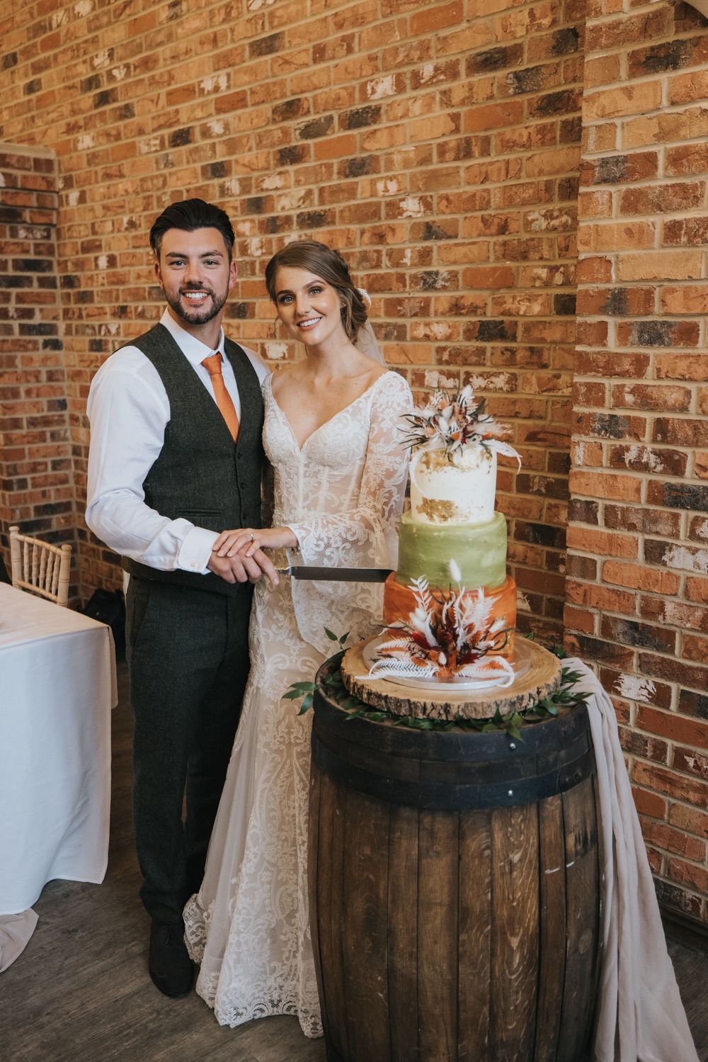 A wedding couple stands beside their rustic two-tier cake displayed on a wooden barrel at Hall Farm - image captured by Grimsby photographer