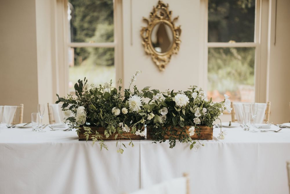 Elegant white floral centerpiece with greenery on long dining table beneath ornate gold mirror at Cleatham Hall.