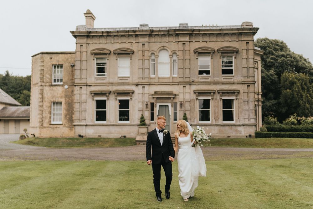 A couple walk hand in hand across a manicured lawn in front of Cleatham Hall on their wedding day.