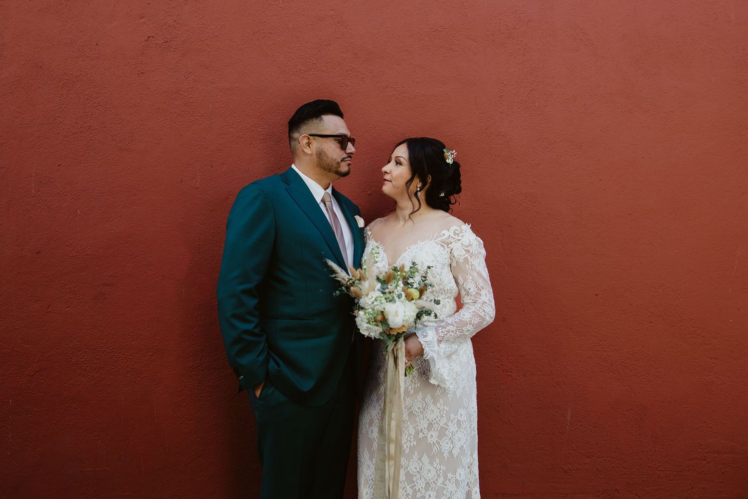 A happy couple shares a sweet moment against a rustic red wall during their wedding celebration.