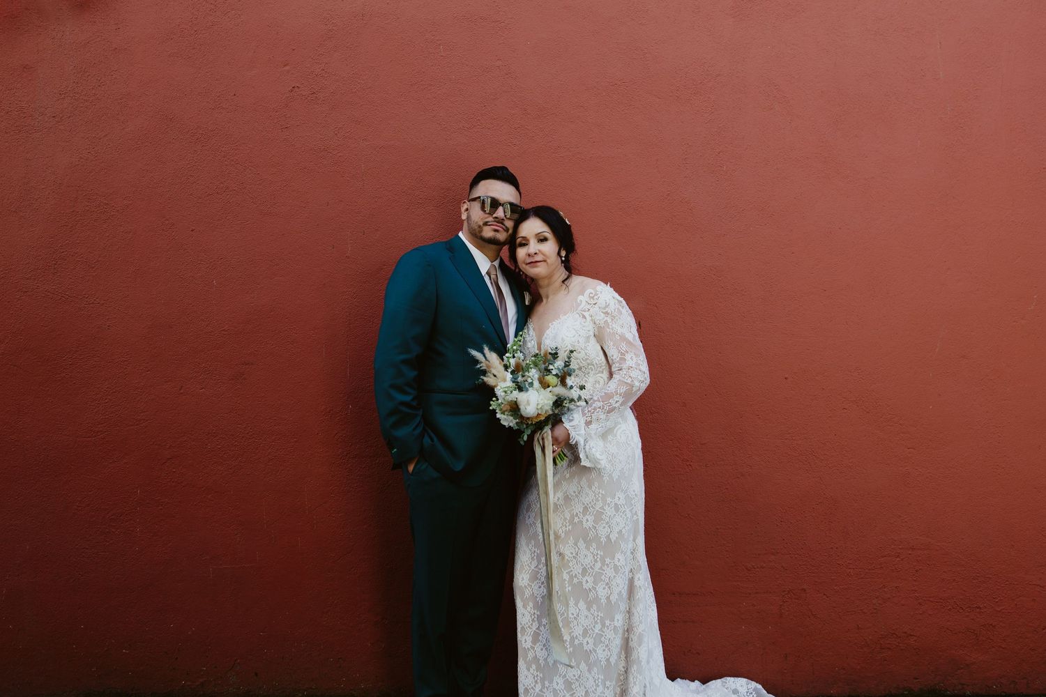Wedding couple poses against a rustic red wall in intimate photo series.