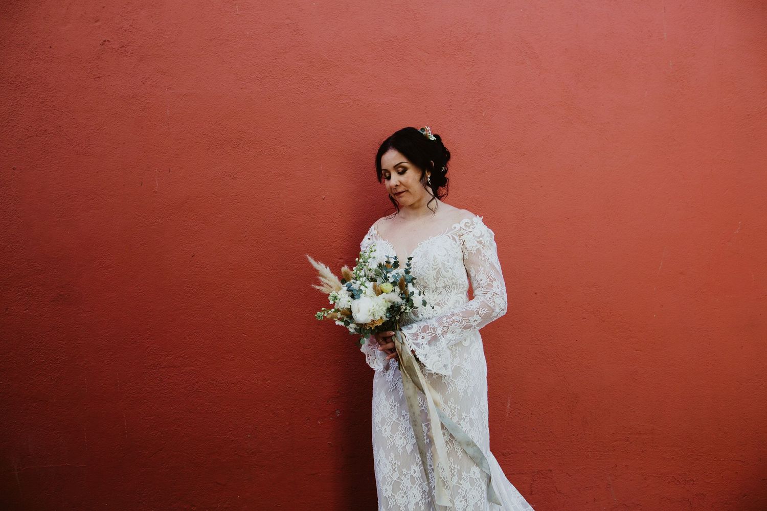 Someone in an ornate lace wedding gown holds a white floral bouquet against a terracotta colored wall.