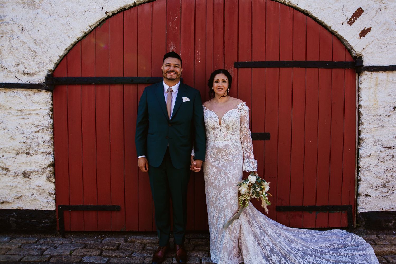 A couple poses in formal wedding attire against a rustic red barn door and white stone wall at their countryside wedding.