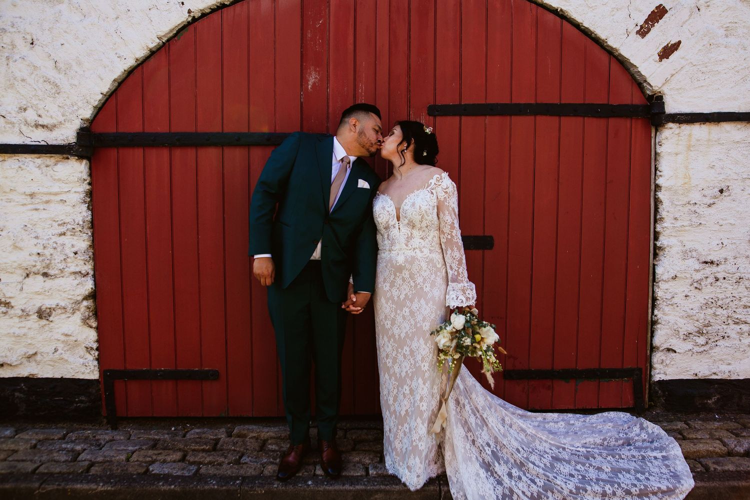 Two people share a romantic moment in front of a rustic red barn door with a bride in a flowing lace gown holding flowers.