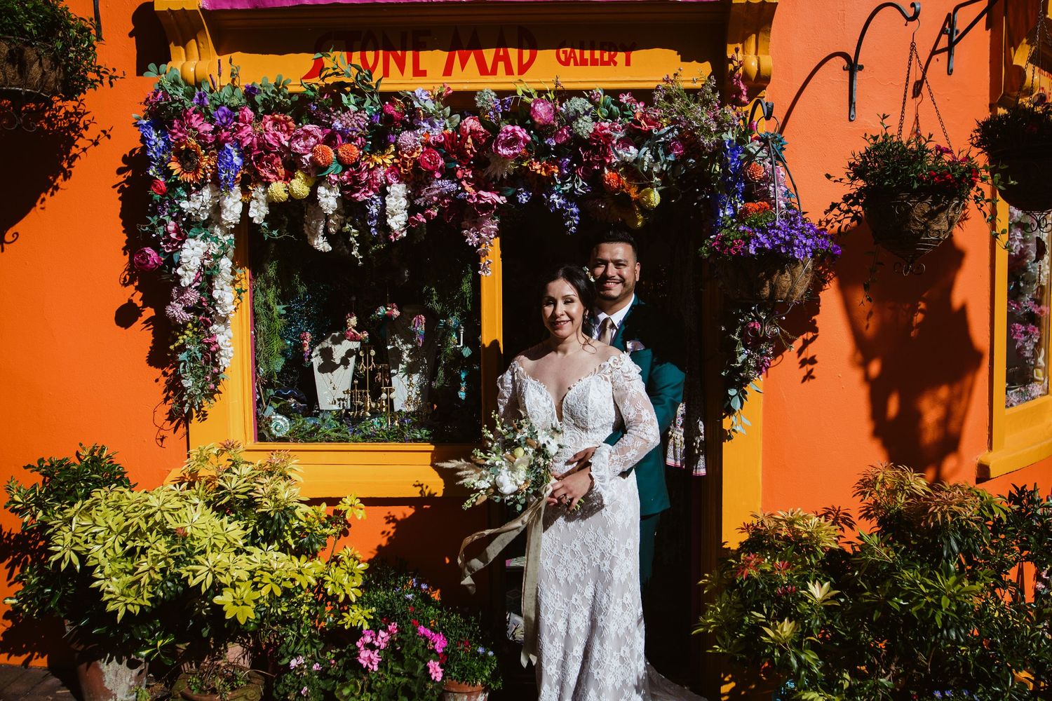 A couple stands beneath a vibrant floral arch against a bright orange wall at a festive venue.