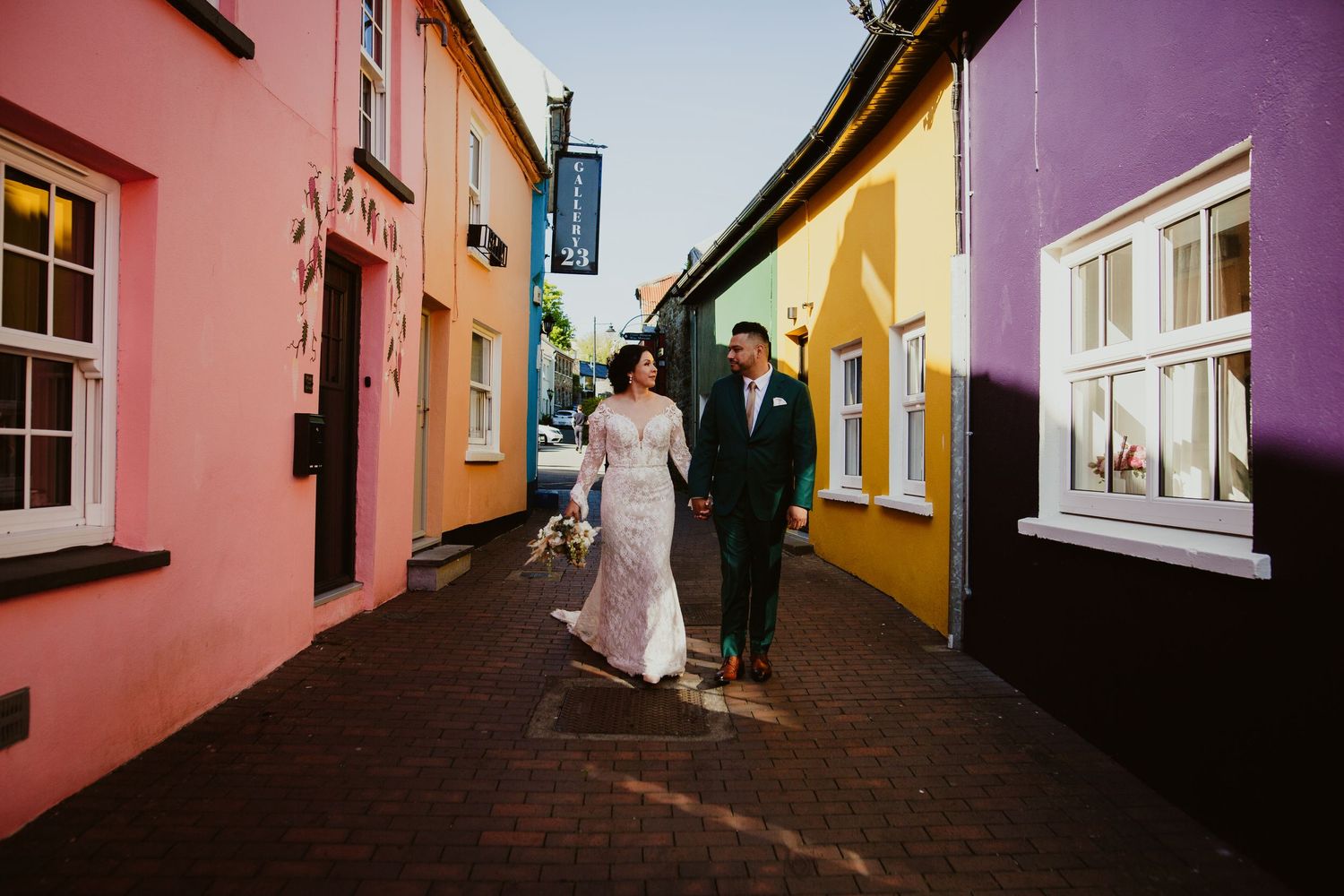 A couple strolls between colorful pink, yellow and purple buildings on a narrow cobblestone street.