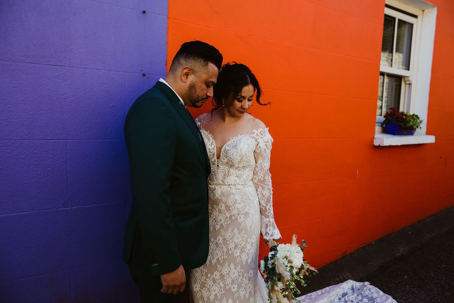 A wedding couple poses against vibrant orange and purple walls in an artistic urban setting.