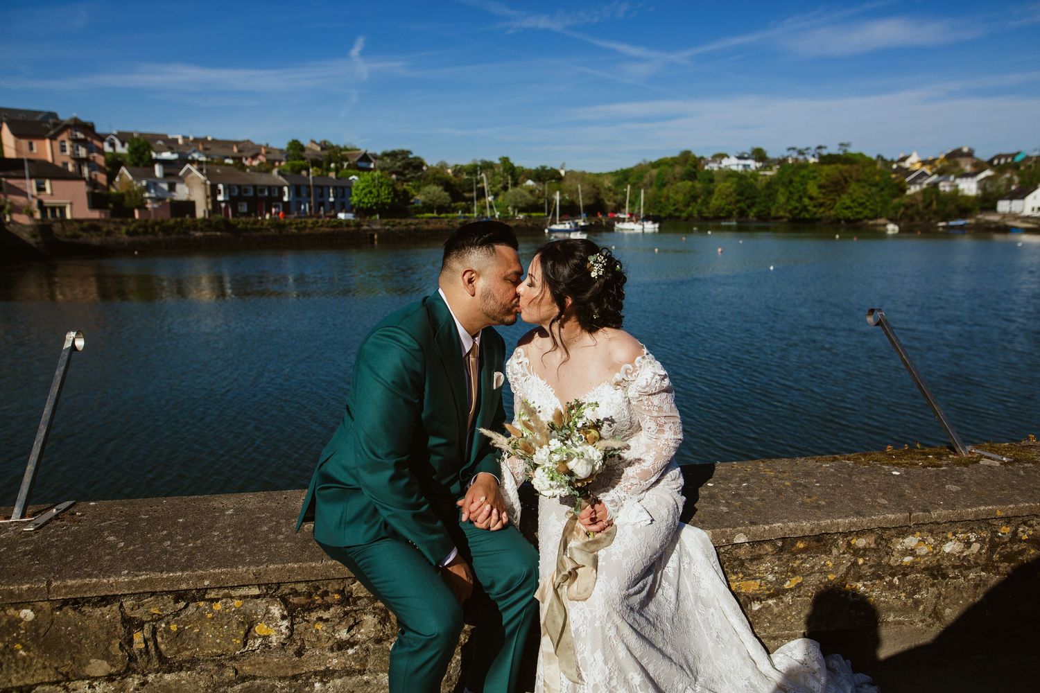 A couple in wedding attire standing along a scenic waterfront harbor with stone walls on a sunny day with blue skies.