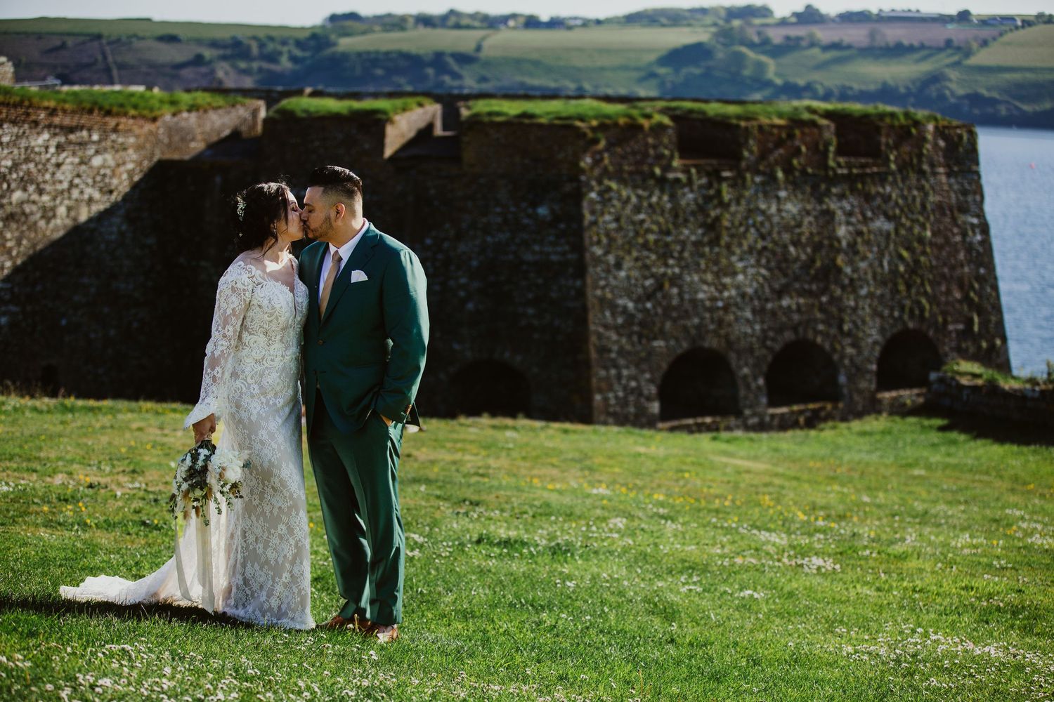 A couple walk hand in hand across lush green coastal cliffs overlooking the ocean on their wedding day.