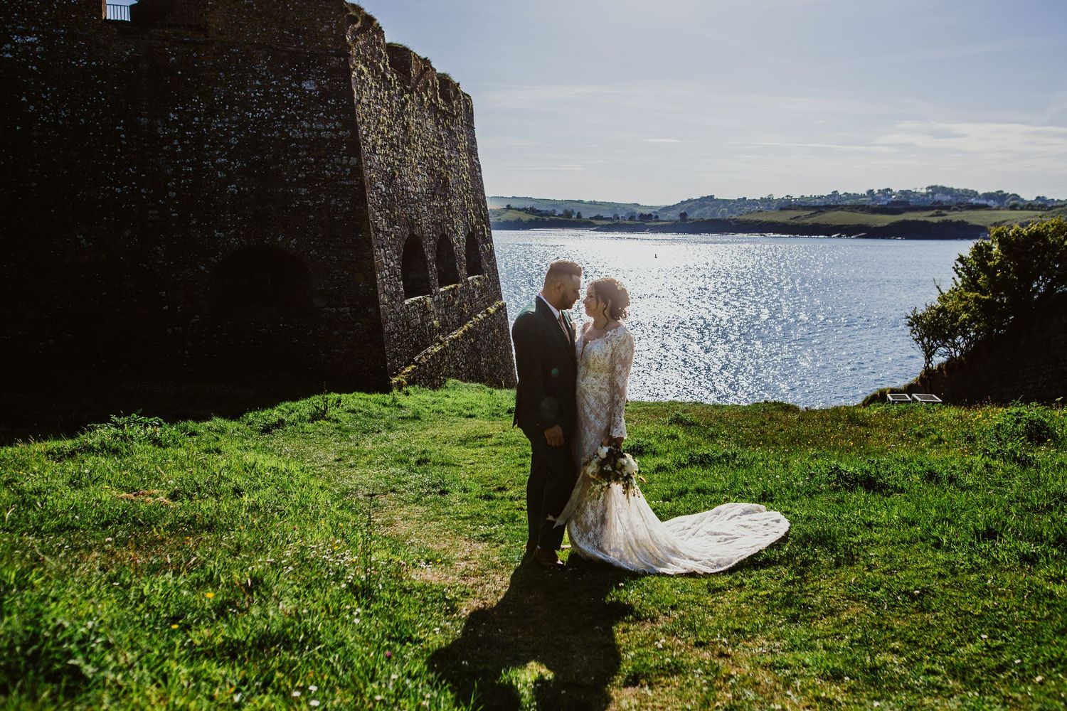 A romantic couple at a coastal stone castle wall overlooking sparkling waters on a sunny day in Ireland.