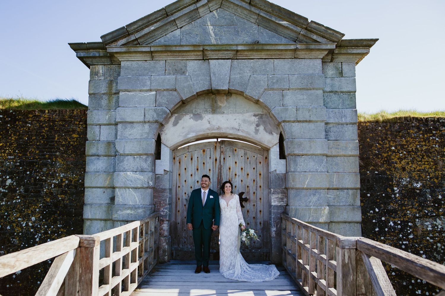 A couple stands in formal attire at the entrance of a historic stone gateway with wooden bridge.
