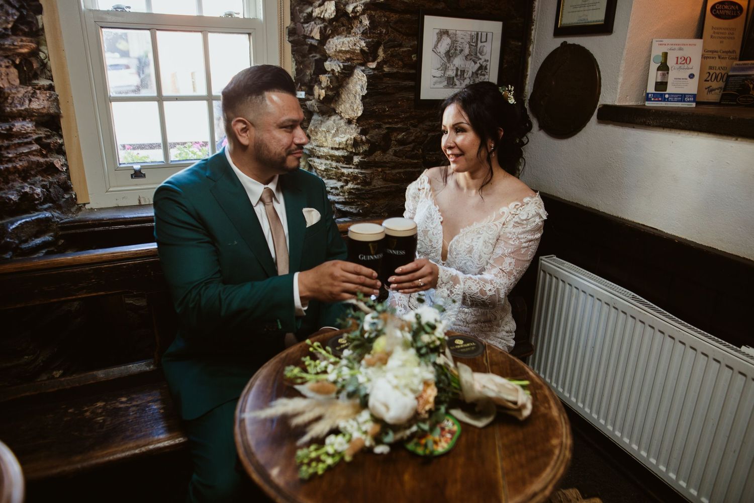 A newlywed couple share an intimate moment at a rustic wooden table inside a cozy stone-walled room.
