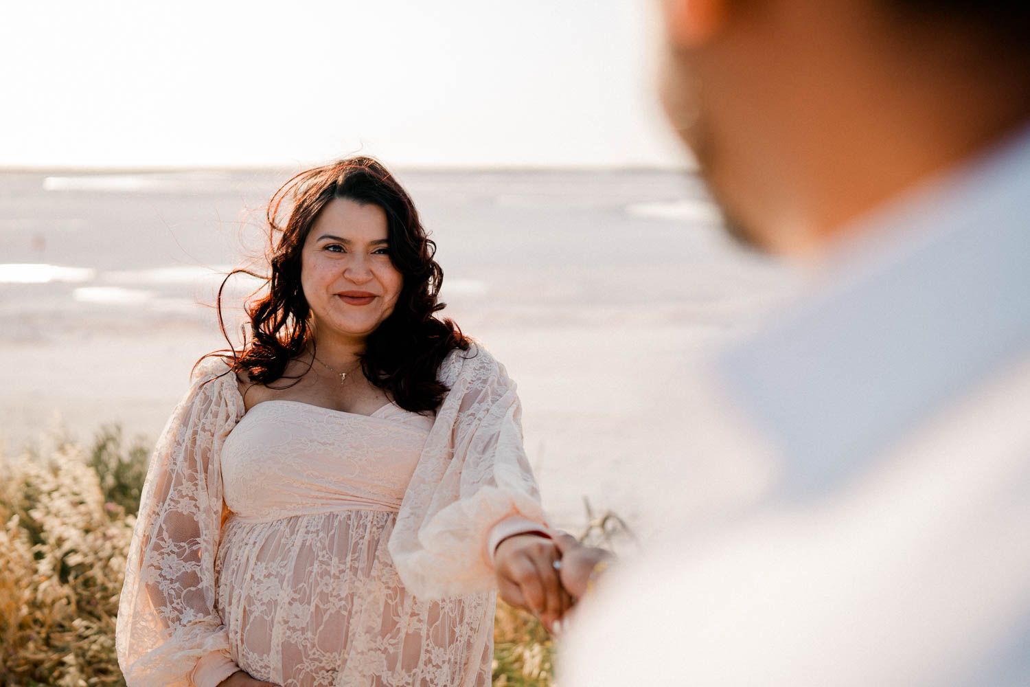 A person in a lace dress and floral shawl stands on a sunny beach during golden hour.