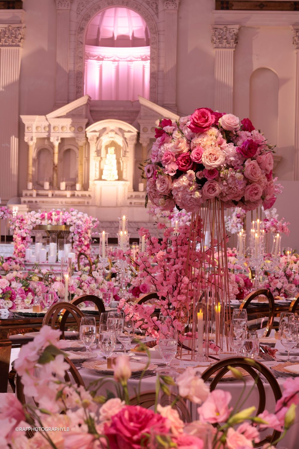 Luxurious pink-themed wedding reception venue with ornate decor, floral centerpieces and elegant table settings bathed in pink lighting.