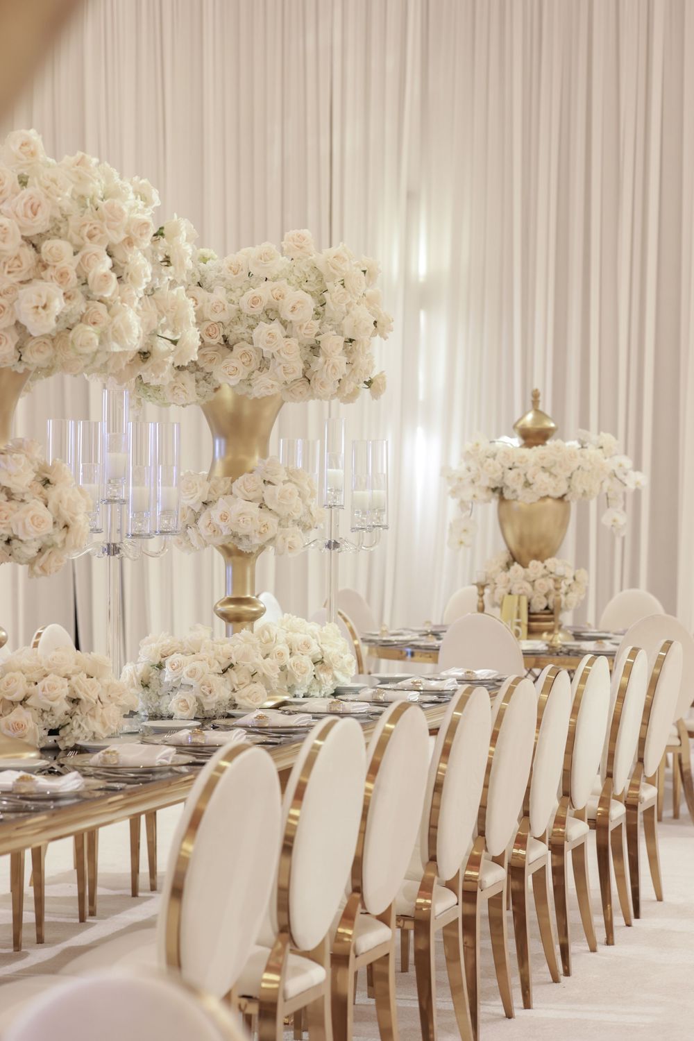 Elegant all-white wedding reception setup with long tables, cream upholstered chairs, and tall floral centerpieces.