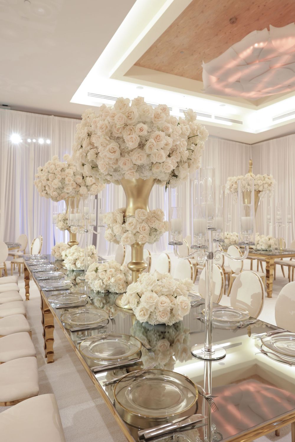 Luxurious white wedding reception table with tall floral centerpieces in gold vases and elegant place settings.