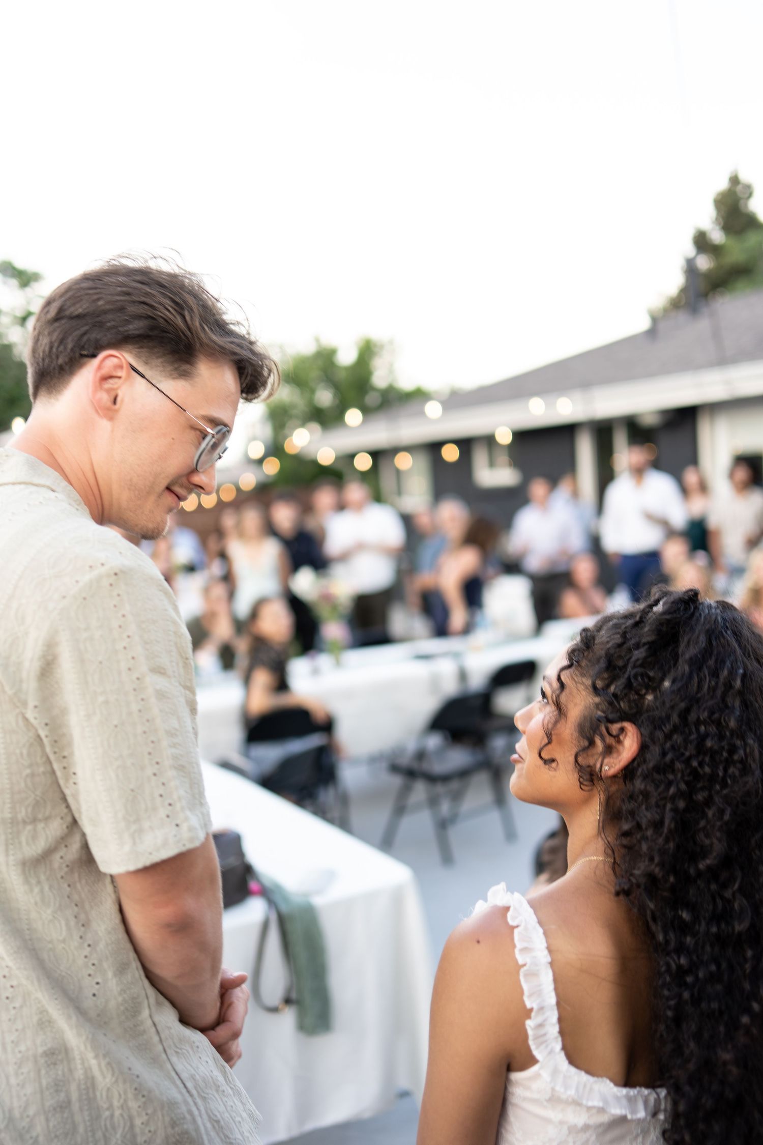 Two people engaged in conversation at an outdoor evening event with string lights and white tables in the background.