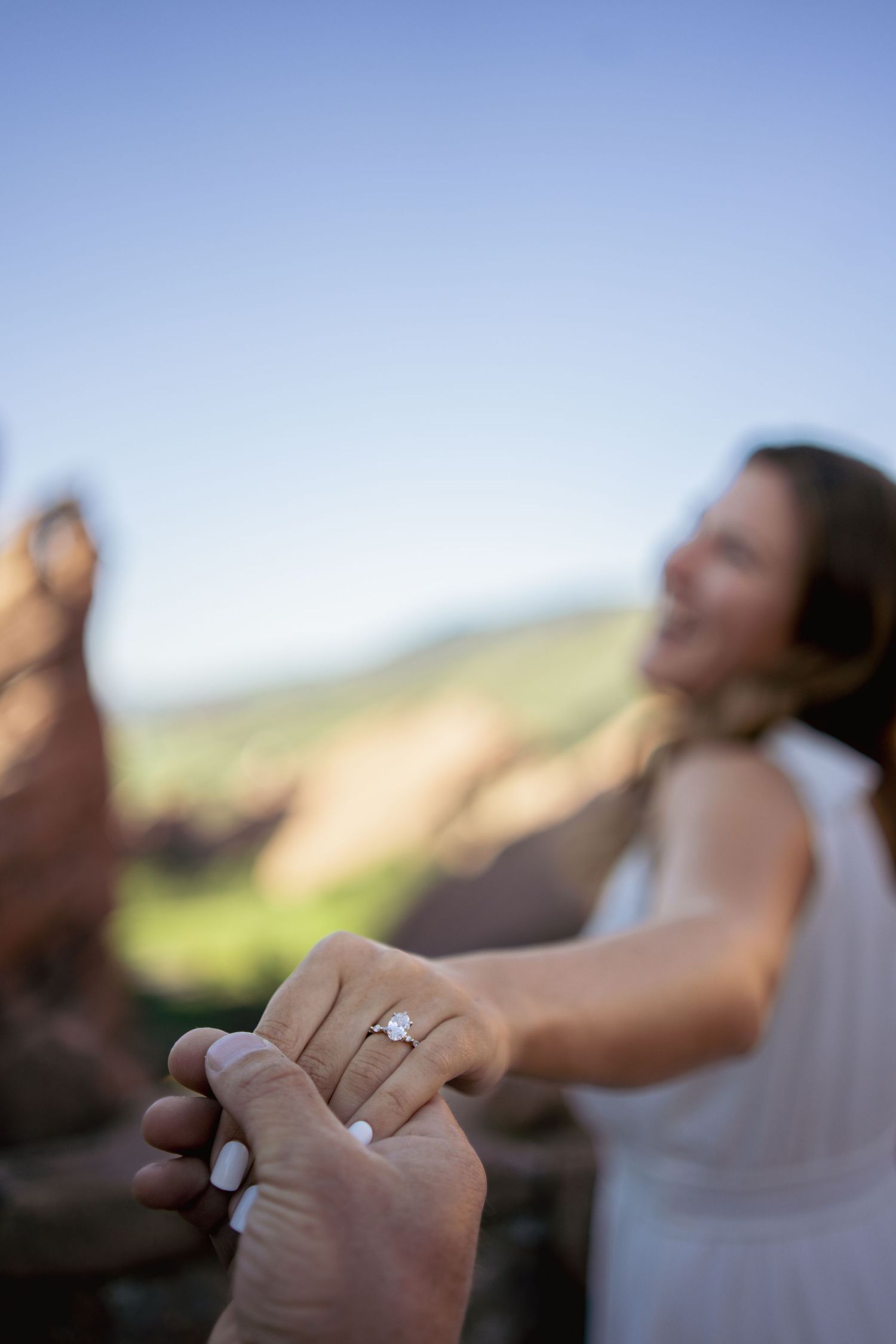 Close-up view of hands holding each other with an engagement ring on display against a blurred outdoor background.