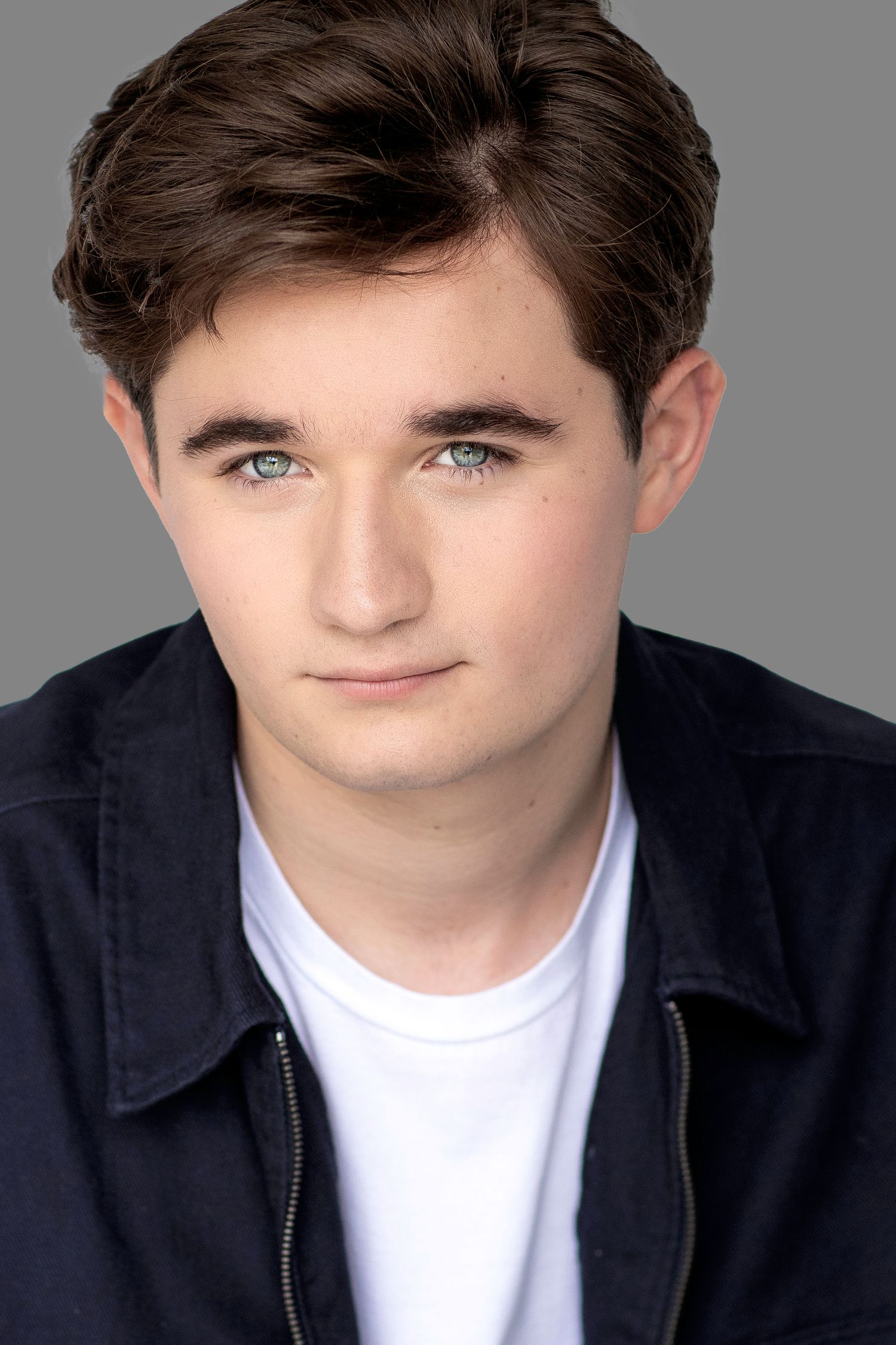 Professional headshot portrait of a young performer in a navy jacket and white t-shirt against a neutral background.