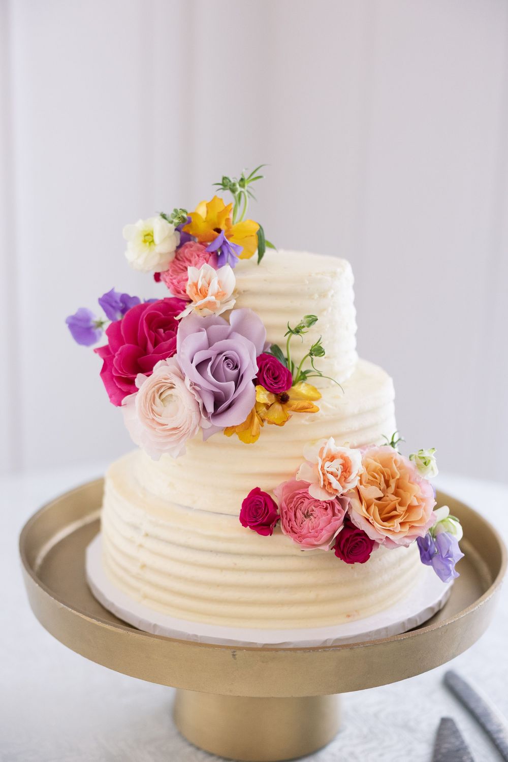 Three-tier white buttercream wedding cake decorated with colorful fresh flowers including roses and pansies on gold stand.