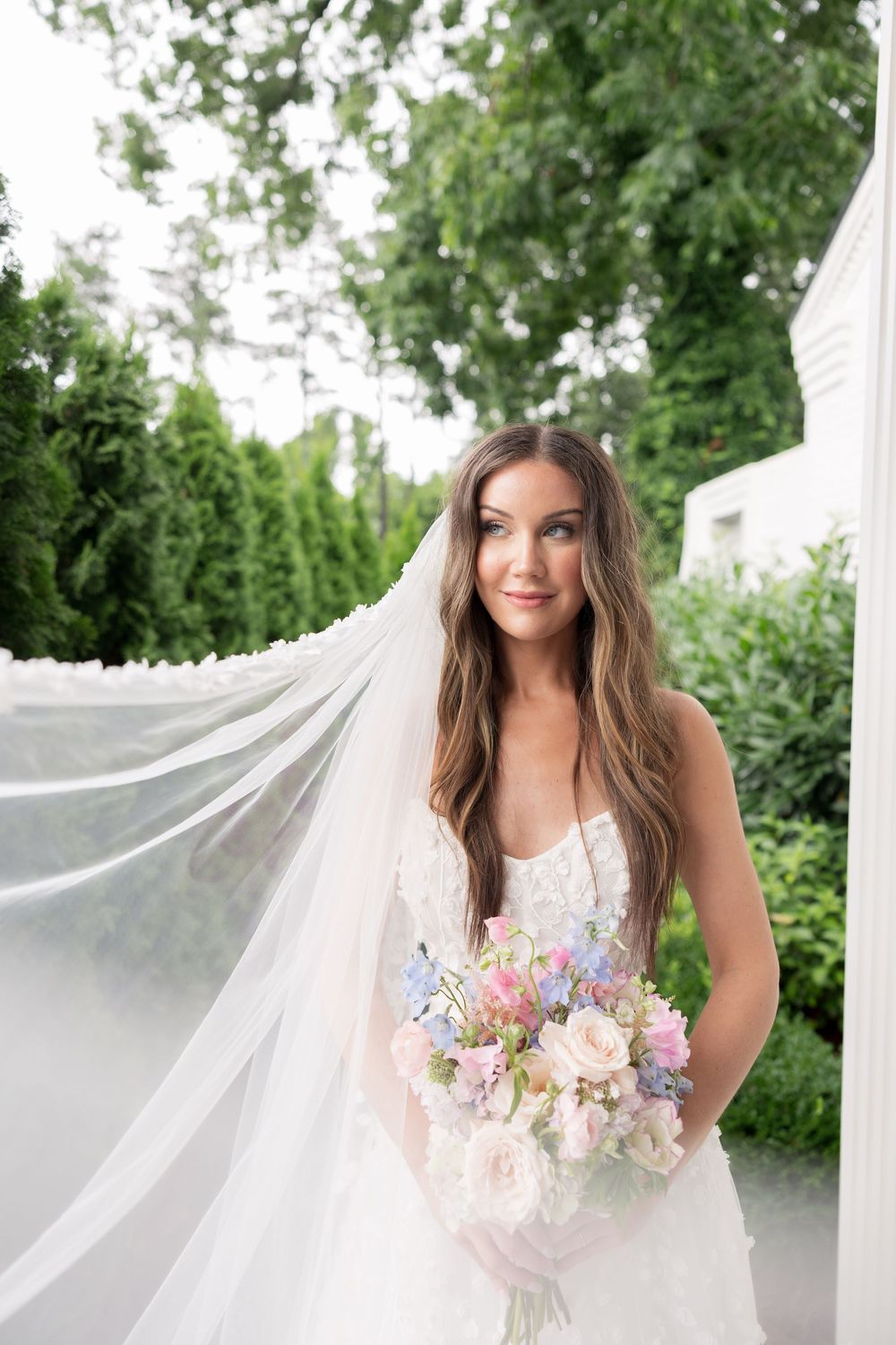 A bride in a white wedding gown holds a pastel bouquet of flowers while her long veil flows in an outdoor garden setting.