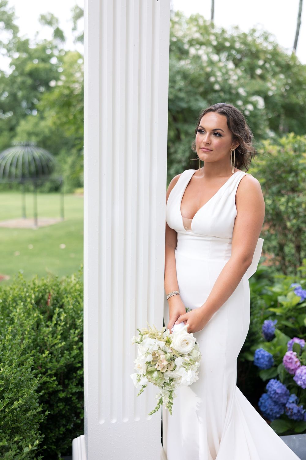 A bride in a fitted white v-neck wedding gown holds a white bouquet while standing beside a classical white column outdoors.