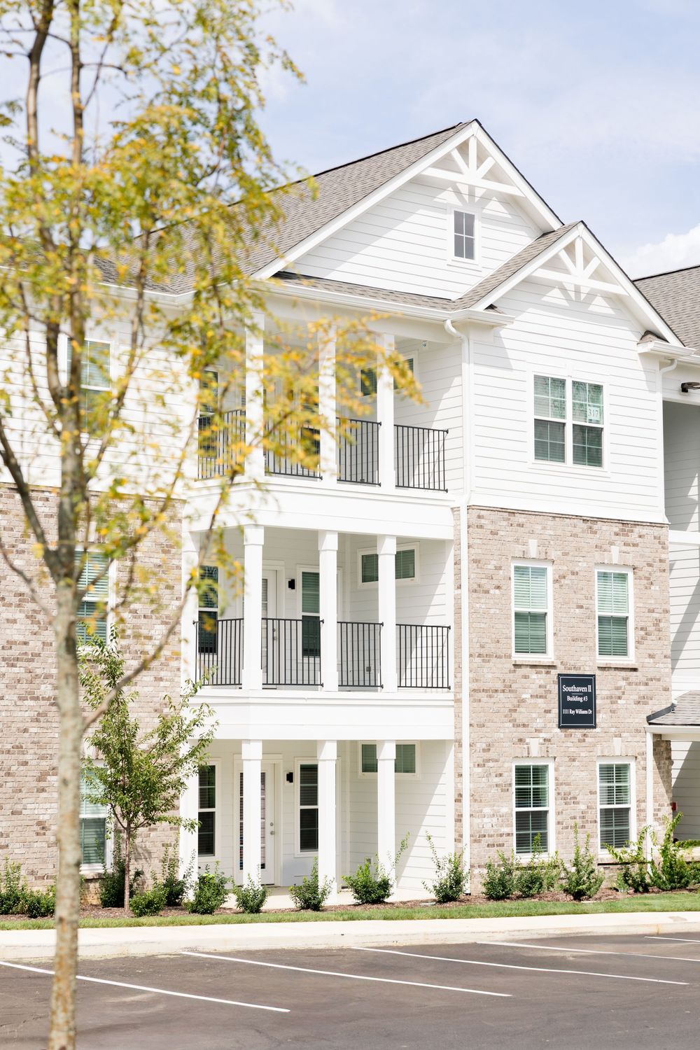 Modern apartment complex exterior featuring white siding, stone accents, and multiple balconies on a tree-lined property.