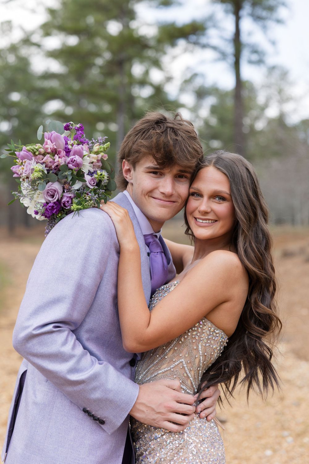 A couple in formal attire pose together outdoors, with a purple and white floral bouquet against a natural background.