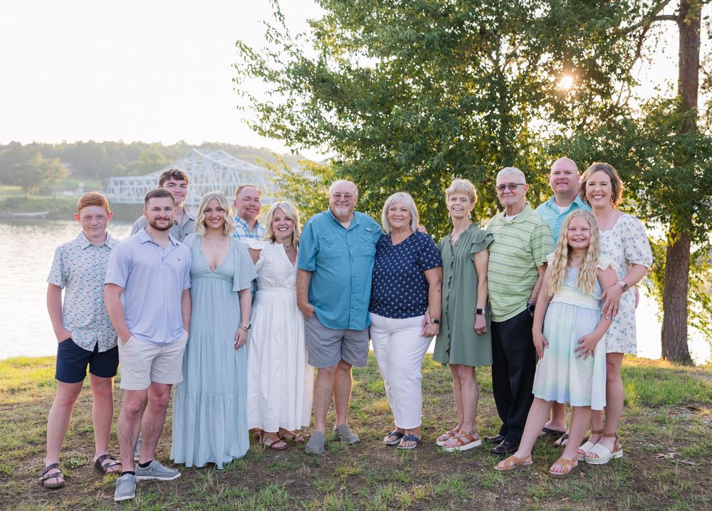 Group wearing pastel and light colored outfits poses together outdoors by a lake during golden hour.