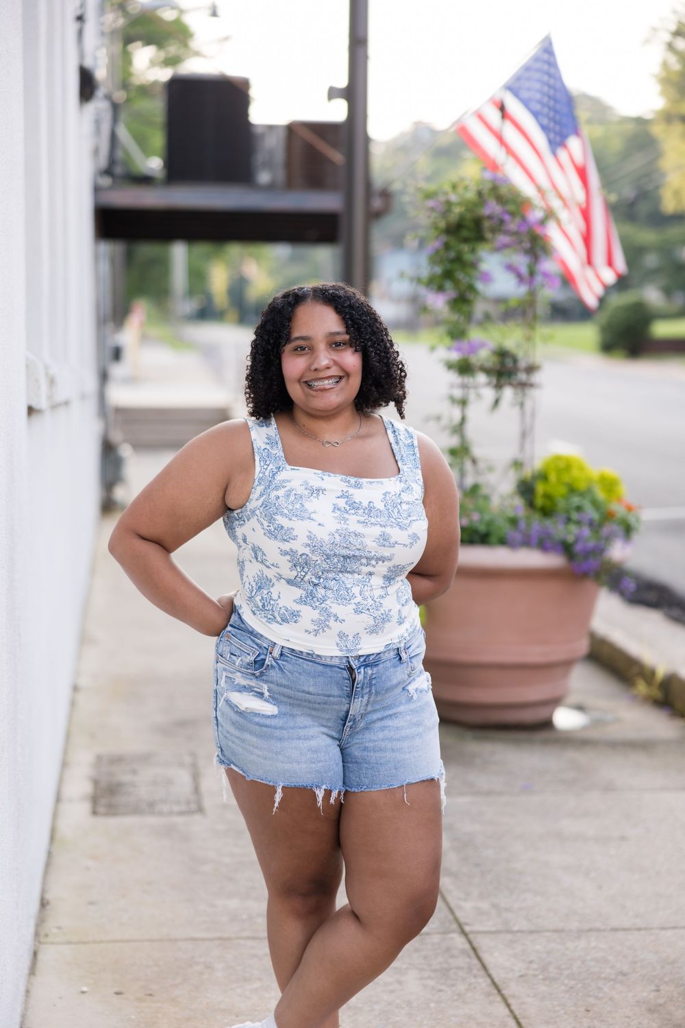 Plus size model in blue floral top and denim shorts poses confidently on porch with American flag in background.