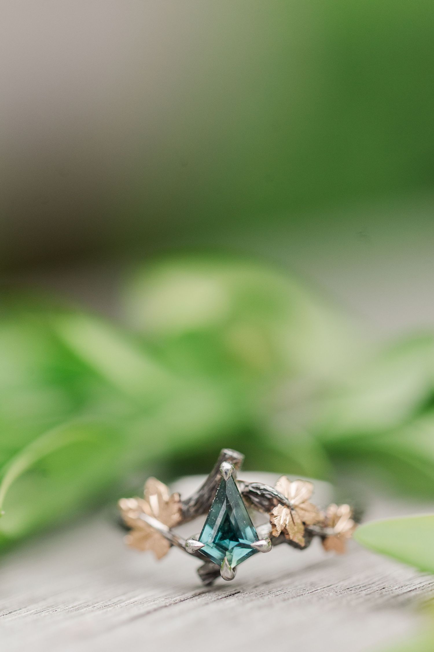 Unique engagement ring with green gemstone and rose gold leaves design displayed on wooden surface with blurred greenery at Cantigny Park