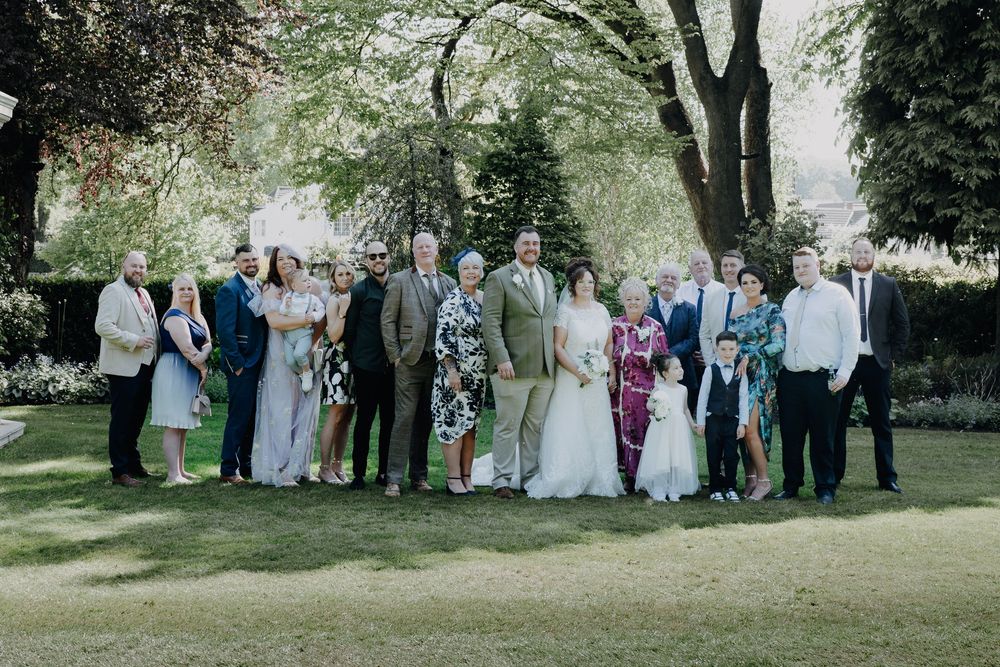 A formal outdoor group photo on a sunny day in a garden with trees and lush greenery in the background.