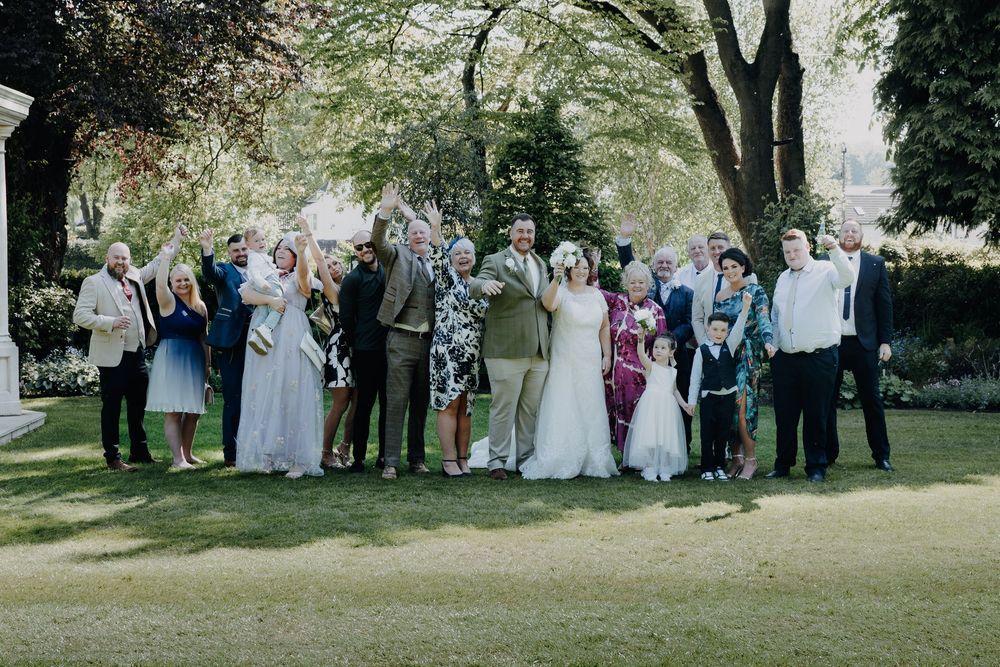 A wedding party gathers for a group photo on a sunny day under green trees in a garden setting.