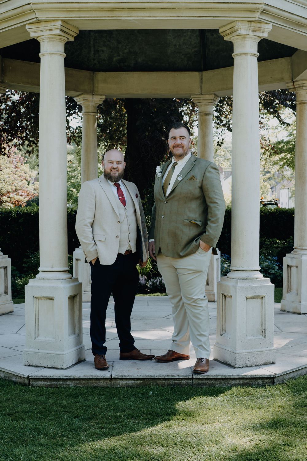 Two people in formal attire stand together in an elegant white gazebo surrounded by landscaped garden grounds.