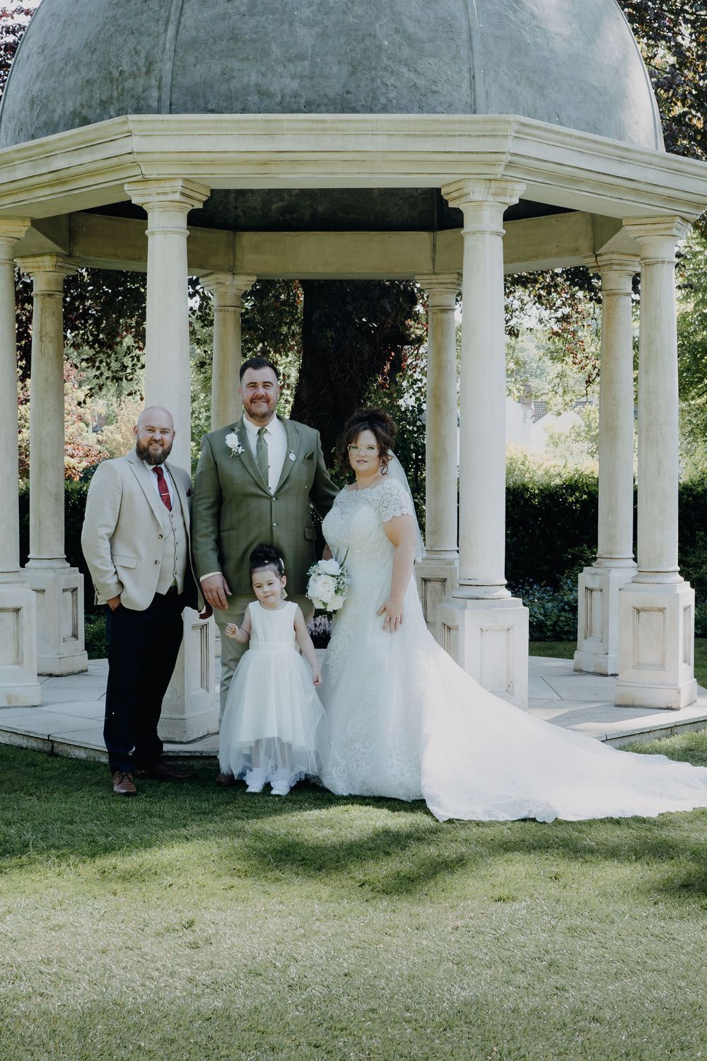 A wedding party poses together under an elegant white gazebo with classical columns on a sunny garden lawn.