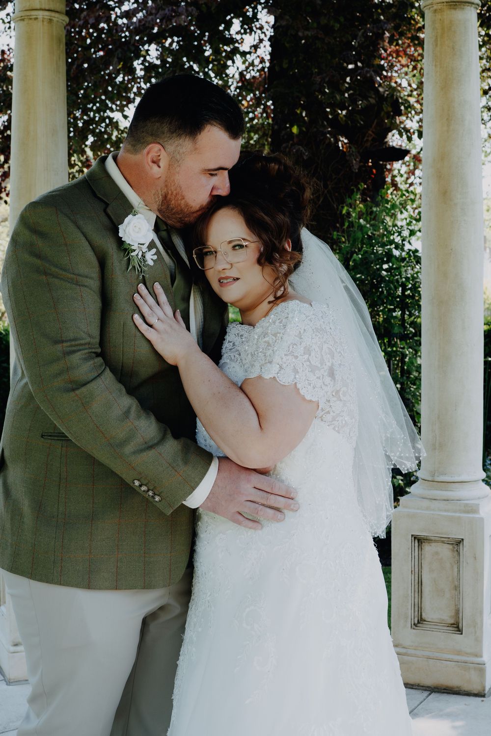 A tender wedding moment captured showing newlyweds embracing on a porch with white columns and greenery in the background.