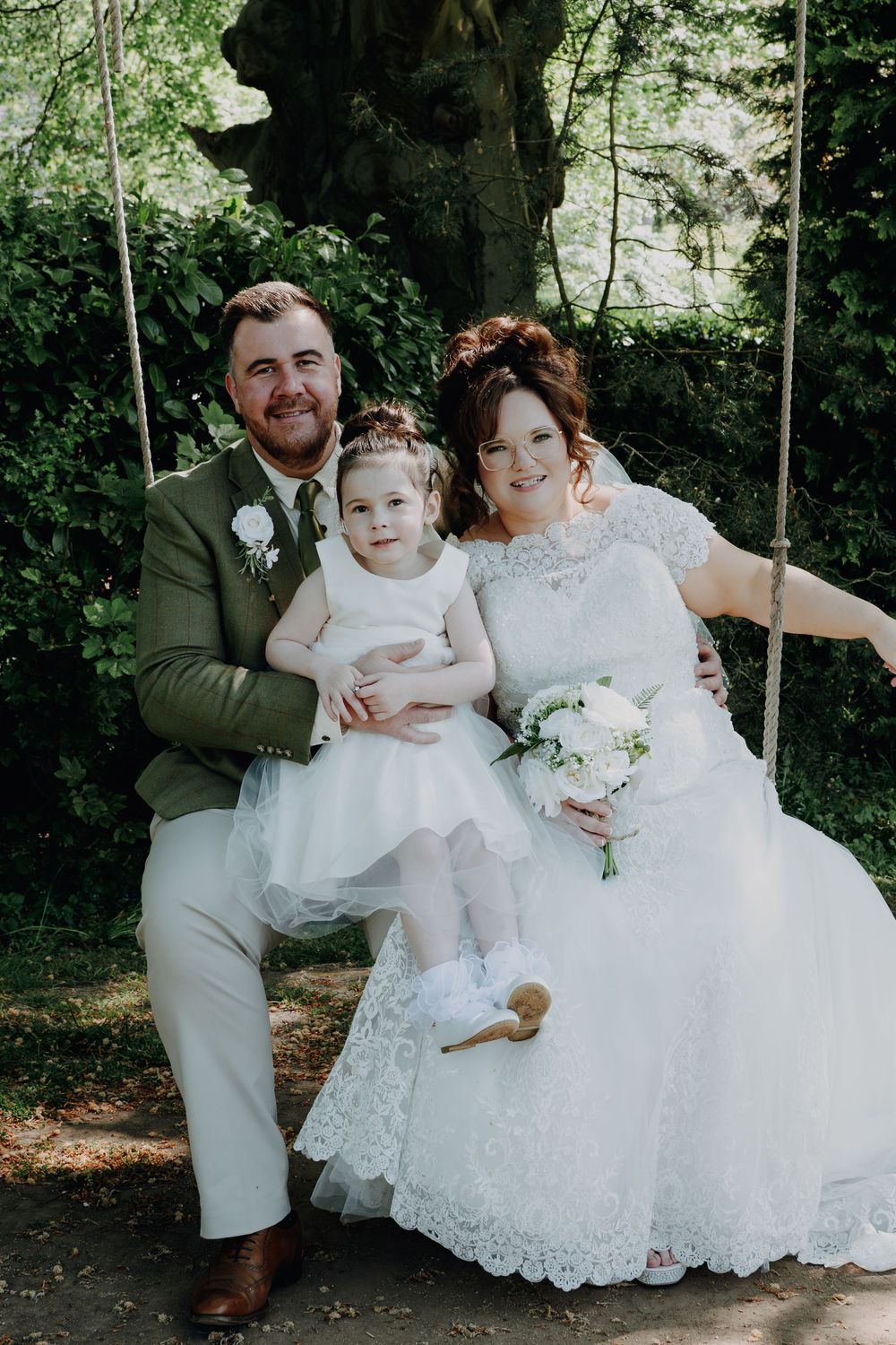 A bride in a lacy white dress sits on a garden swing next to her loved ones during an outdoor wedding photo.