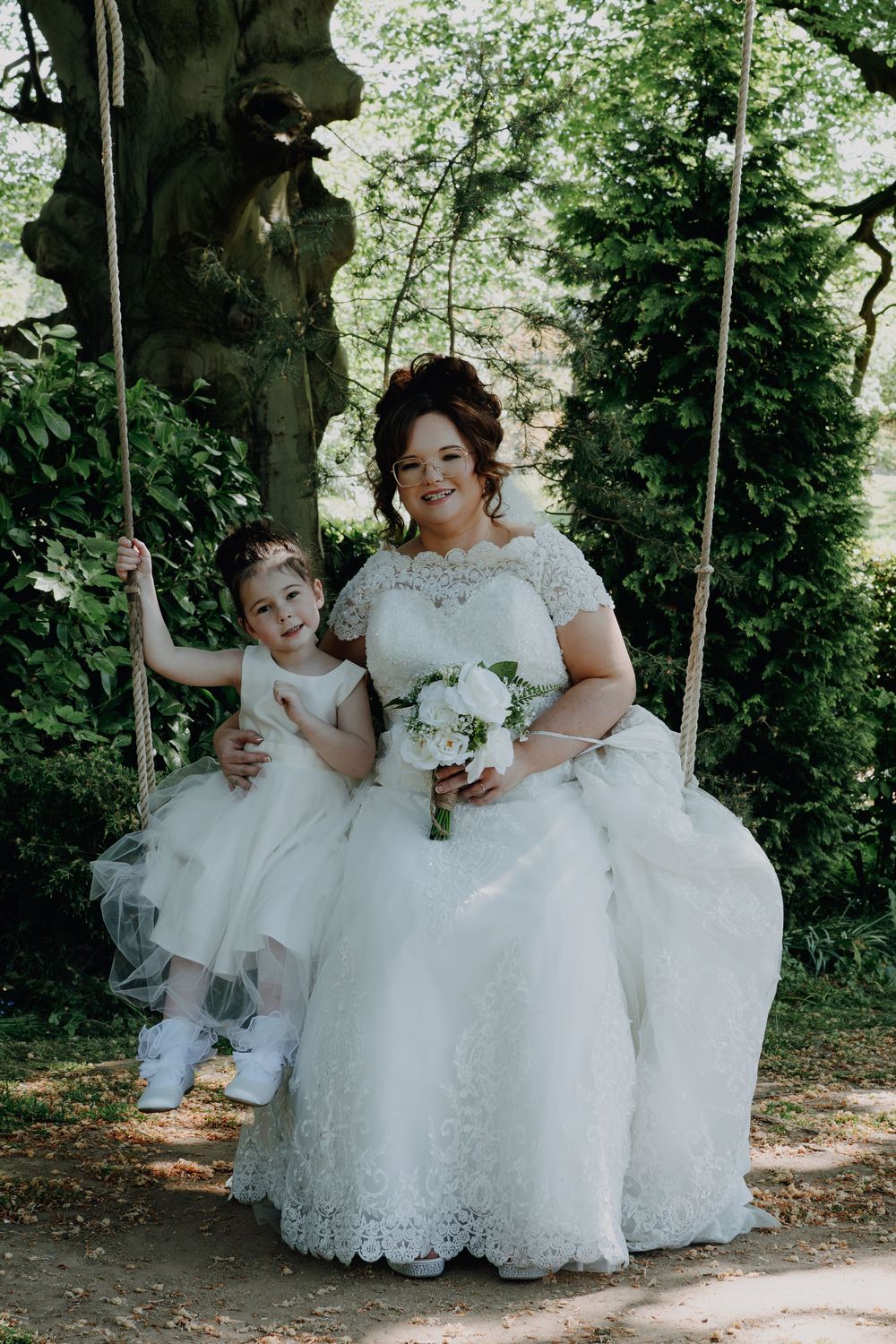 A bride in a white lace gown and young child in white dress sit together on a wooden swing in a garden setting.