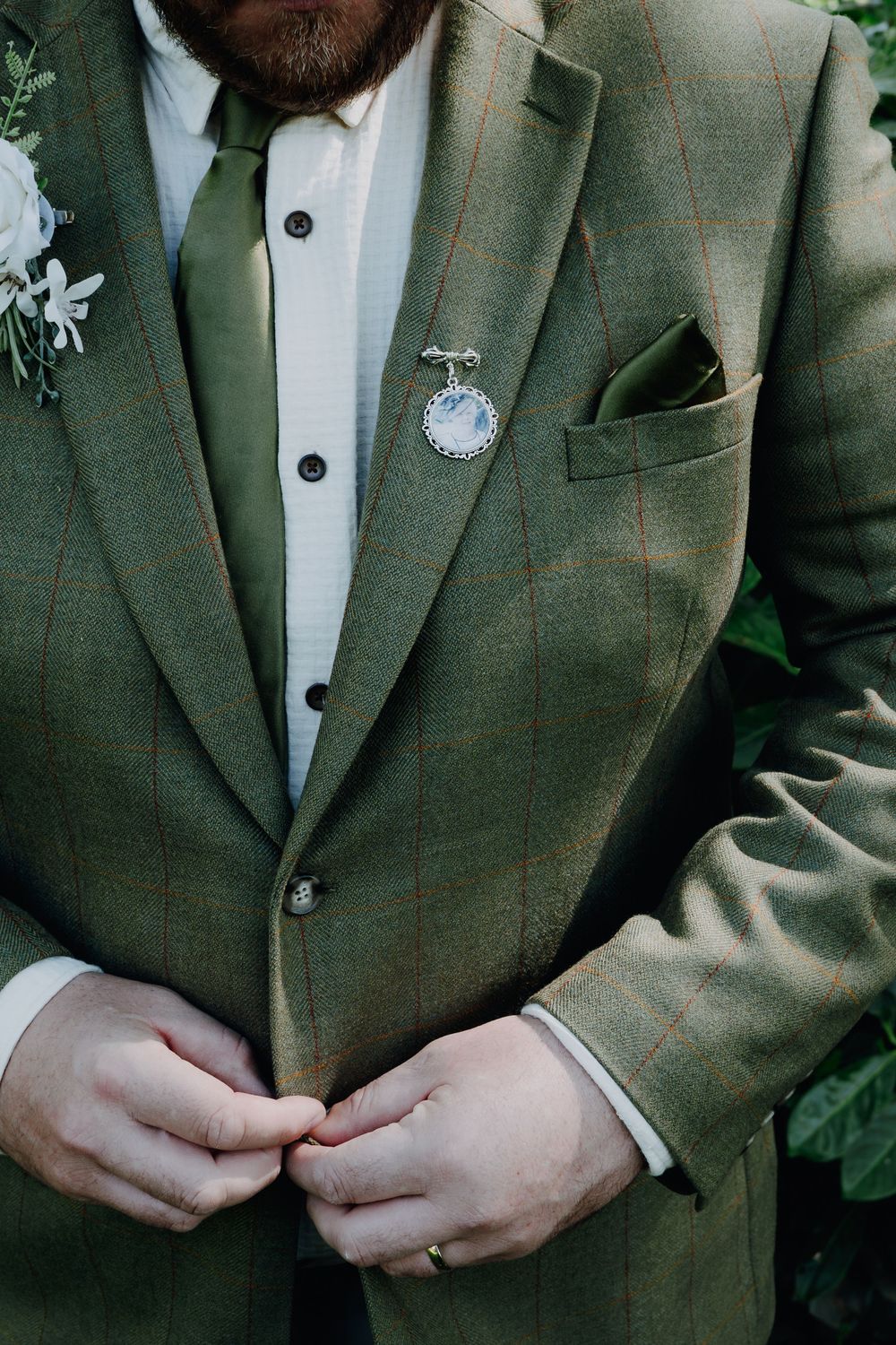 A close-up of a green suit jacket with a white boutonniere and pocket watch accessory against a leafy background.