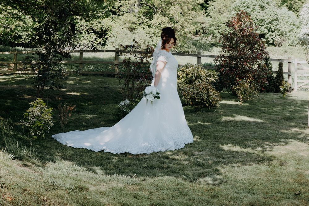 A bride in a flowing white lace wedding dress stands in a sunlit garden surrounded by greenery and flowers.