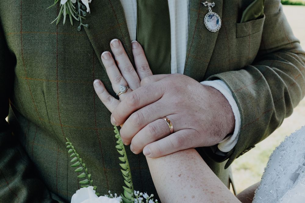 Close-up detail of hands wearing wedding rings with green suit jacket and white floral boutonniere in view.