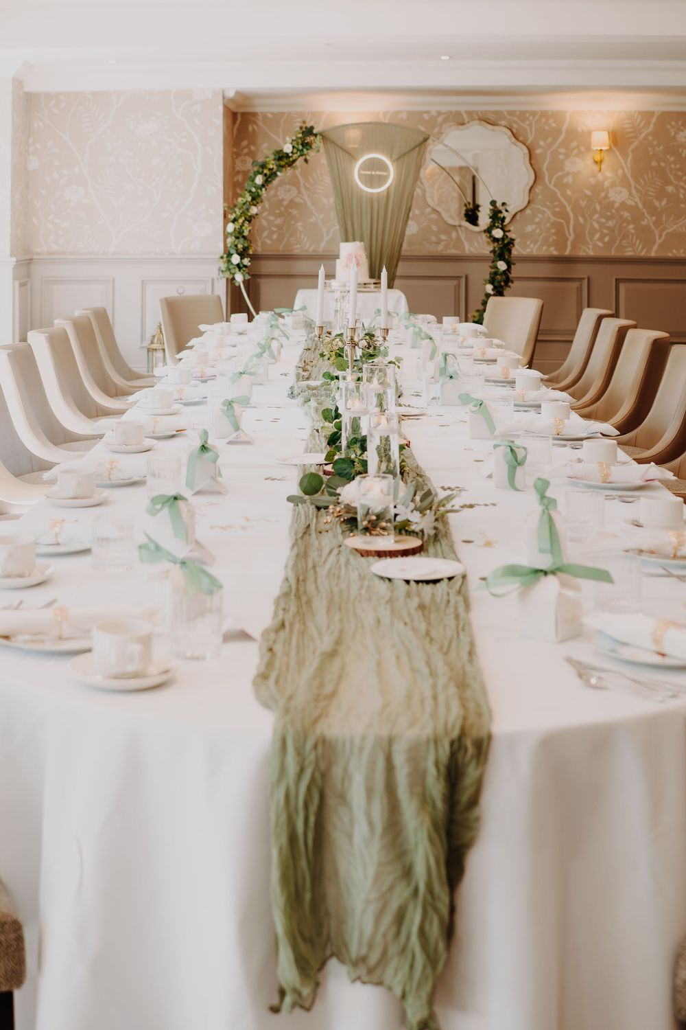 An elegant wedding reception table set in white with sage green accents and floral centerpieces.