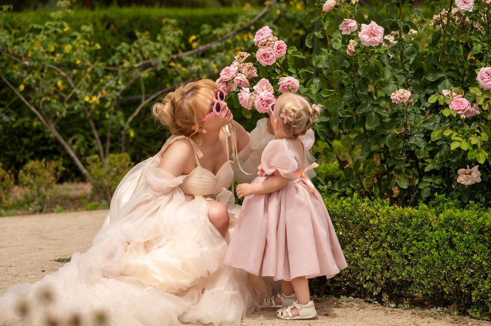 Two figures in light pink dresses share a tender moment in a blooming rose garden on a sunny day.
