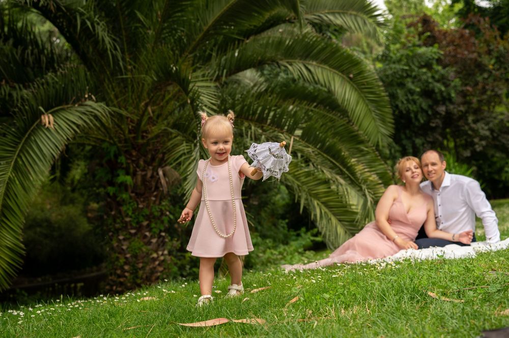 Toddler plays with flowers on grassy hill while couple rests on blanket near tropical palm trees in garden setting.