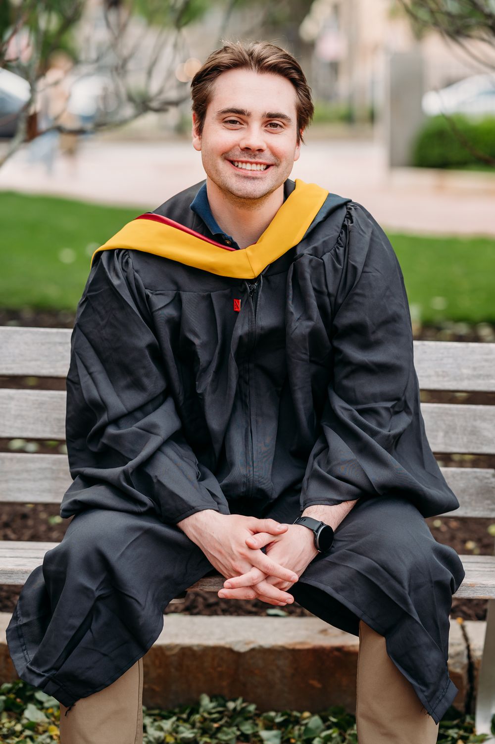Graduate in black cap and gown with yellow hood sits smiling on wooden bench outdoors during commencement.
