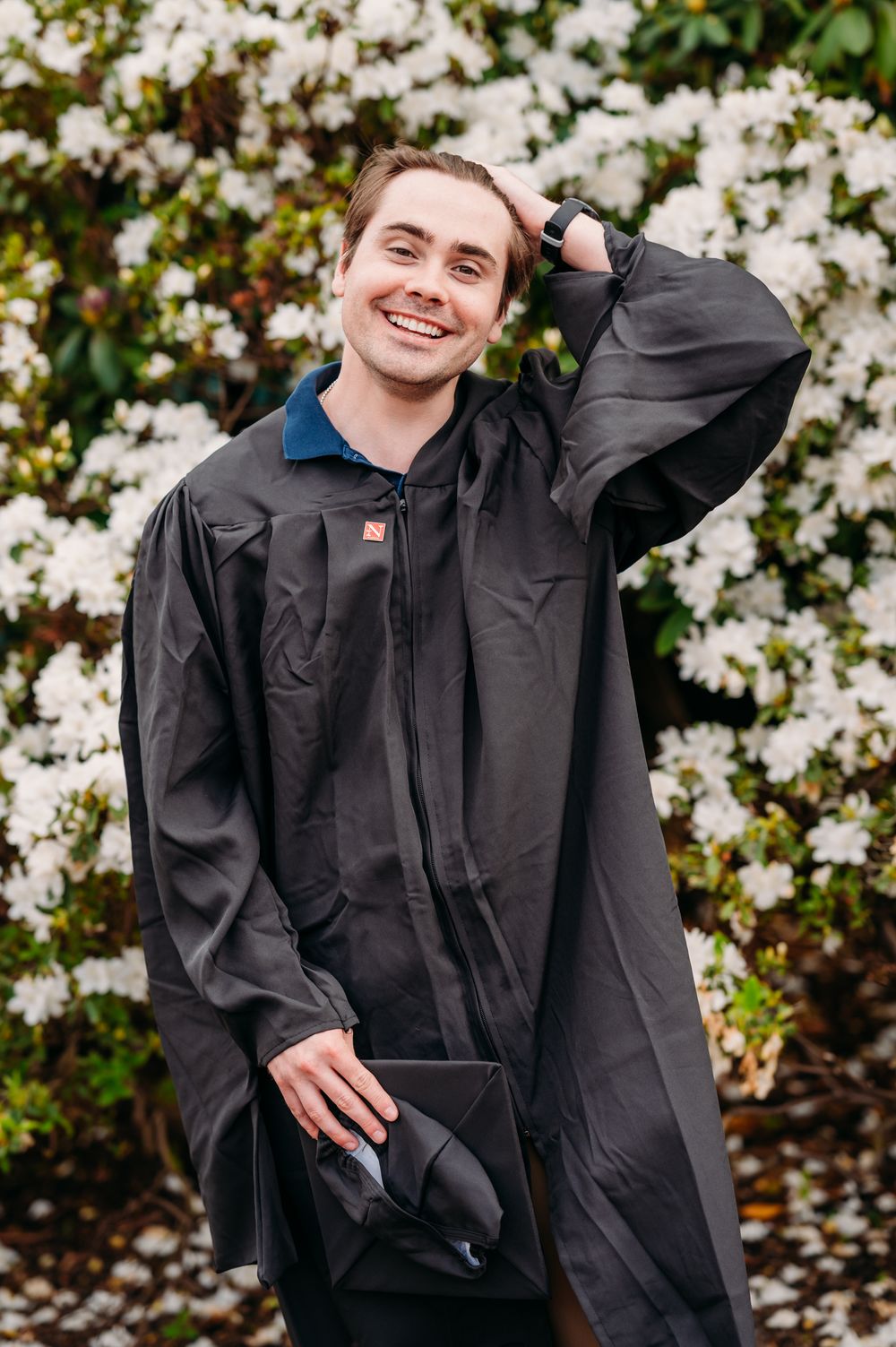 Someone wearing a black raincoat stands smiling in front of blooming white spring flowers in a garden.