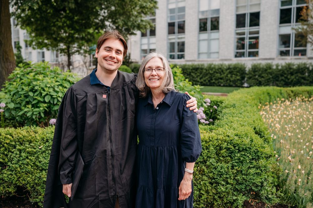 A graduation photo in a garden setting with two people standing together by hedges and a building.